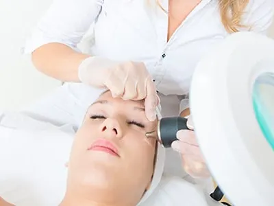 A clinician in gloves using a handheld device on a woman's face beside a magnifying lamp