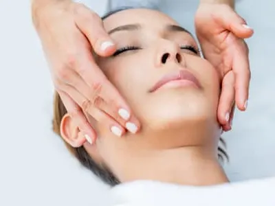 A woman lies with eyes closed as two hands rest on either side of her face during a facial