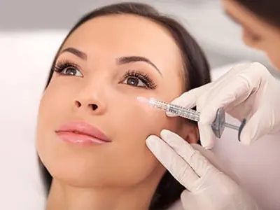 A clinician in gloves holds a syringe to a woman's cheek near her eye