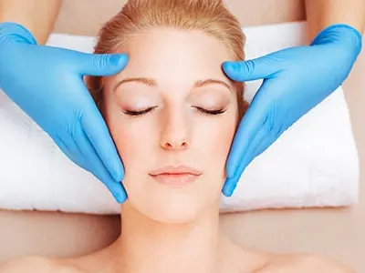 A woman lies on a white towel with eyes closed as hands in blue gloves rest at her temples
