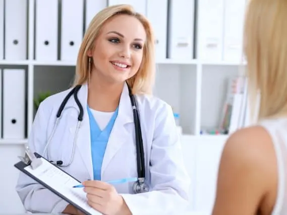 A smiling doctor in a white coat with a stethoscope holds a clipboard while talking with a woman.