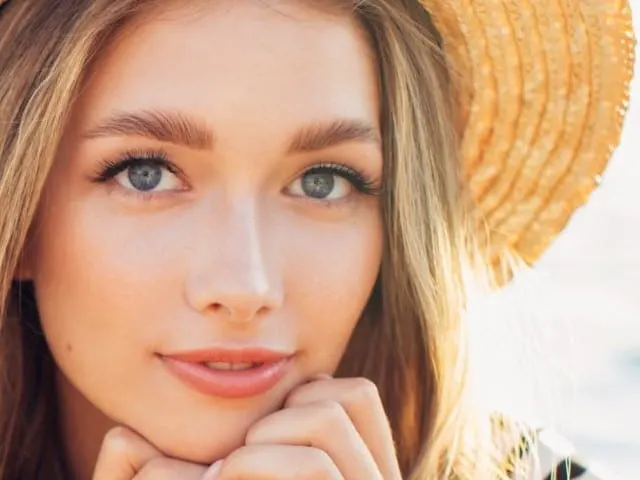 A woman in a straw hat rests her chin on her hands with the sea behind her.
