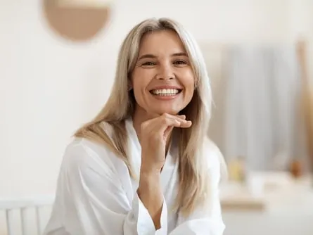 A smiling blonde woman in a white robe rests her chin on her hand.