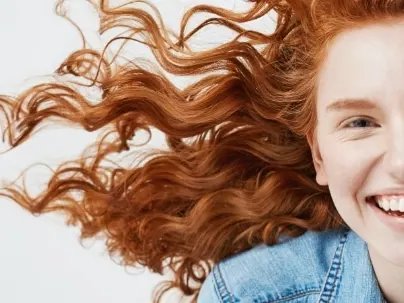 A smiling young woman with curly red hair blowing around her face, wearing a denim jacket