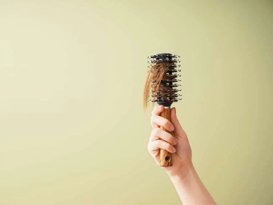 A hand holds a round hairbrush with strands of brown hair caught in the bristles.