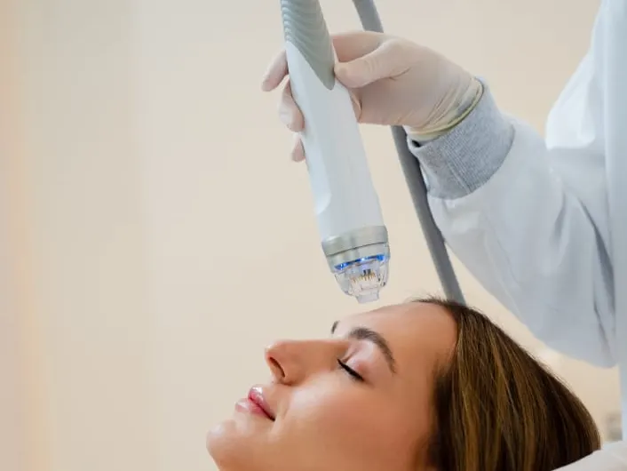 A model lies back with her eyes closed while a gloved clinician holds a handheld device over her face