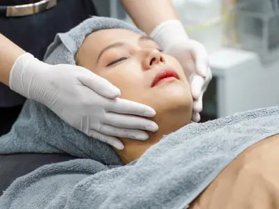 A model reclines under a towel with her eyes closed while a gloved esthetician holds her face during a facial