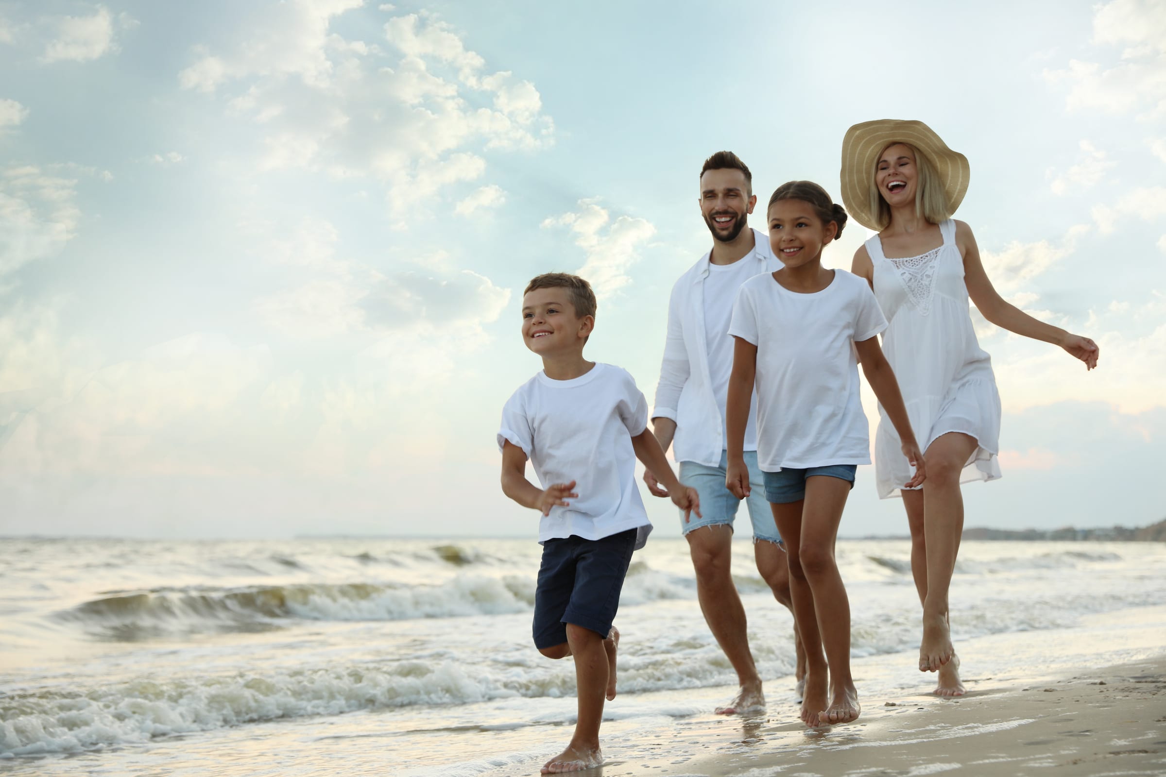 Models: a smiling mother in a sun hat, a father, a daughter and a son running together along a sandy beach by the sea