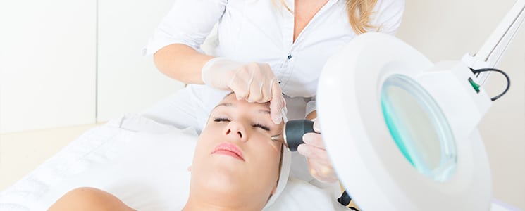 A clinician in gloves using a handheld device on a woman's face beside a magnifying lamp