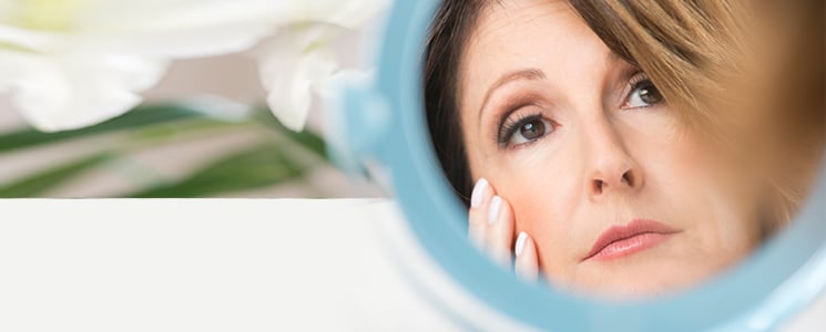 A mature woman looks at her reflection in a small hand mirror, one hand touching her cheek