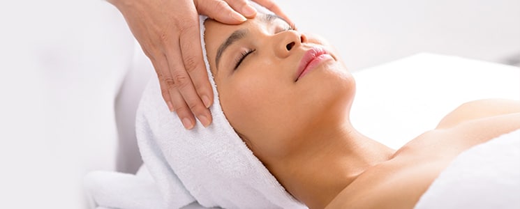 A woman with her hair in a white towel lies back with eyes closed as hands press on her forehead