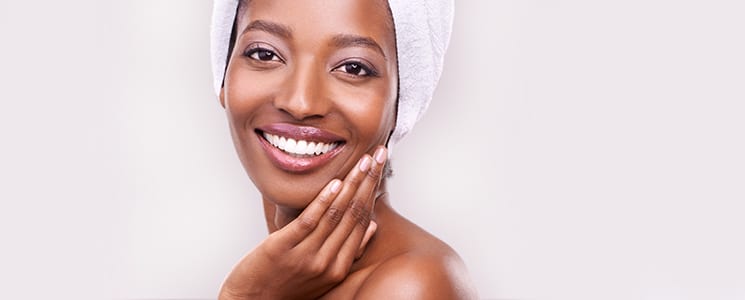 A smiling woman with her hair wrapped in a white towel rests her hand against her cheek