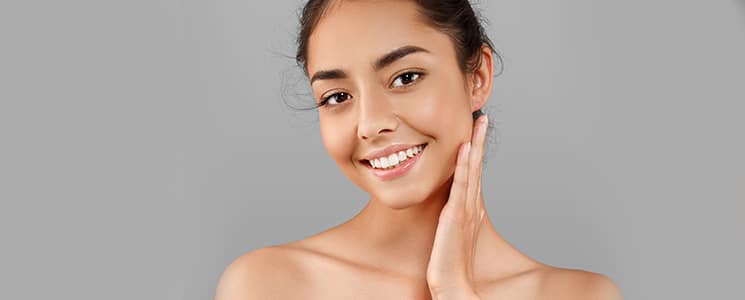 A smiling young woman with bare shoulders touches her neck against a gray background