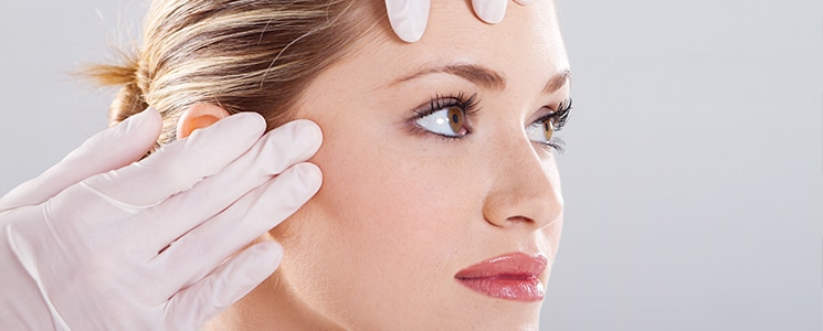 A woman looks to the side as a gloved hand touches her temple and forehead