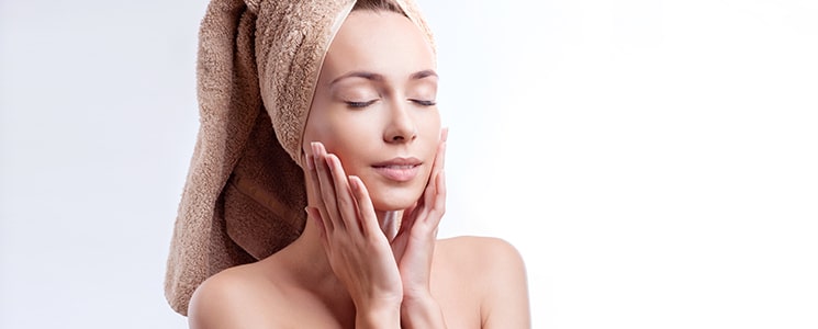 A woman with her hair in a brown towel and eyes closed touches her cheeks with both hands