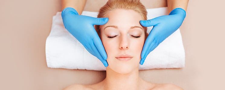 A woman lies on a white towel with eyes closed as hands in blue gloves rest at her temples