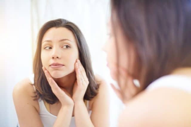 A woman with brown hair touches her cheeks while looking at her face in a mirror.