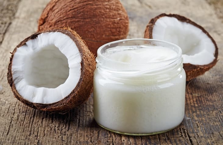 A jar of white coconut oil beside two halved coconuts on a wooden table.