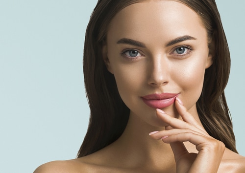 A woman with long brown hair and blue eyes touches her lip with her fingertips.
