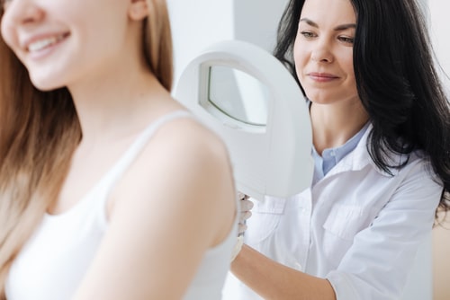 A dermatologist holds a lighted magnifier near a young woman's shoulder in a clinic.