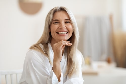 A smiling blonde woman in a white robe rests her chin on her hand.