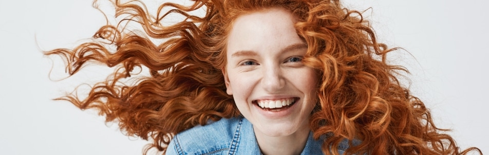 A smiling young woman with curly red hair blowing around her face, wearing a denim jacket