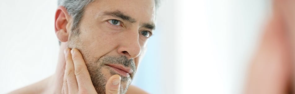 A man with gray hair and stubble touching his jaw while looking at his reflection
