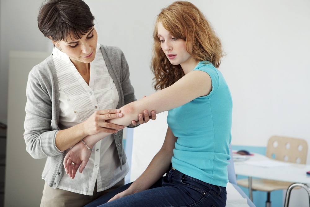 A woman in a gray cardigan examines the red skin on a young woman's elbow in an exam room.