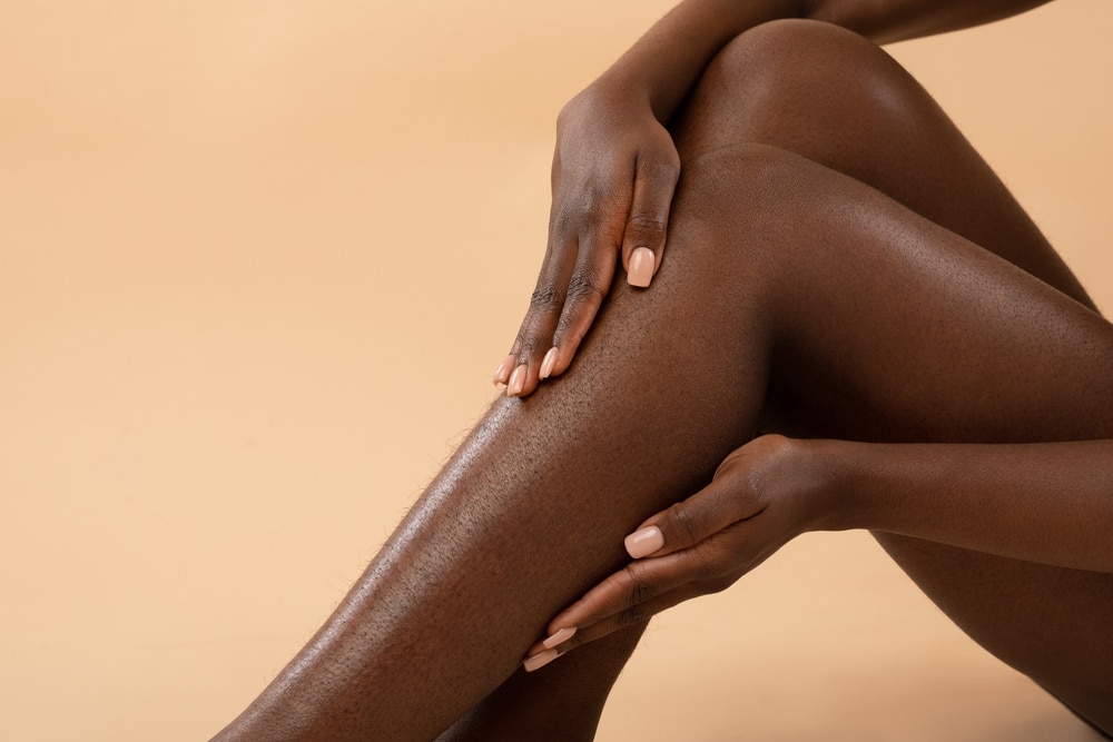 A woman's bare legs, her hands resting on her shin and knee, against a tan background.