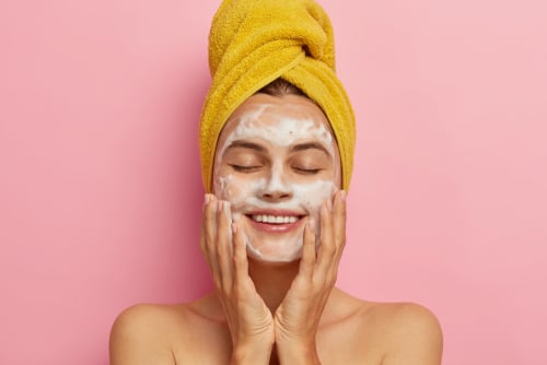 A smiling woman in a yellow head towel washes her face with white foam against a pink background.