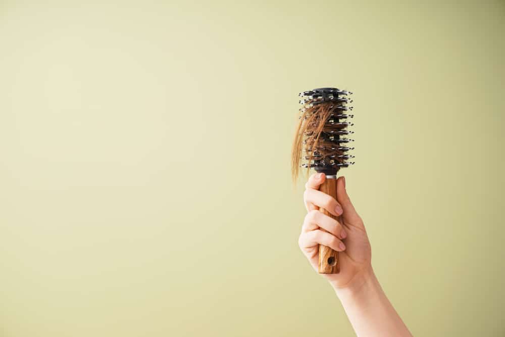 A hand holds a round hairbrush with strands of brown hair caught in the bristles.