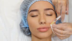 A model in a surgical cap lies with her eyes closed as a gloved clinician injects filler into her lips