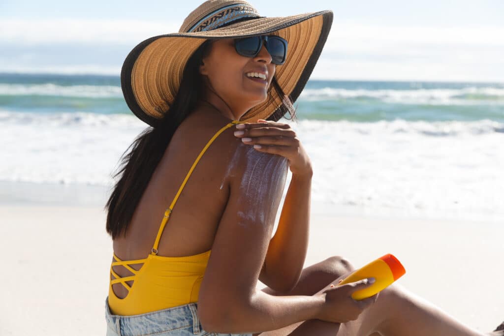 Woman on beach applying sunscreen to shoulders to protect against melanoma and skin cancer.
