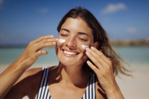 Woman applying sunscreen to her face at the beach to protect her skin from sun exposure.
